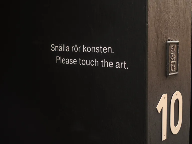 The gallery's black entrance at Svartensgatan 10 with the text: Snälla rör konsten. Please touch the art.