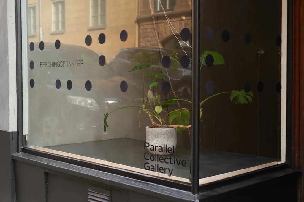 The gallery window with the exhibition title Beröringspunkter, enlarged braille dots on the glass and a monstera in a concrete pot.