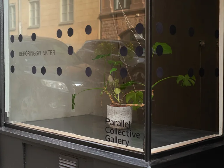 The gallery window with the exhibition title Beröringspunkter, enlarged braille dots on the glass and a monstera in a concrete pot.