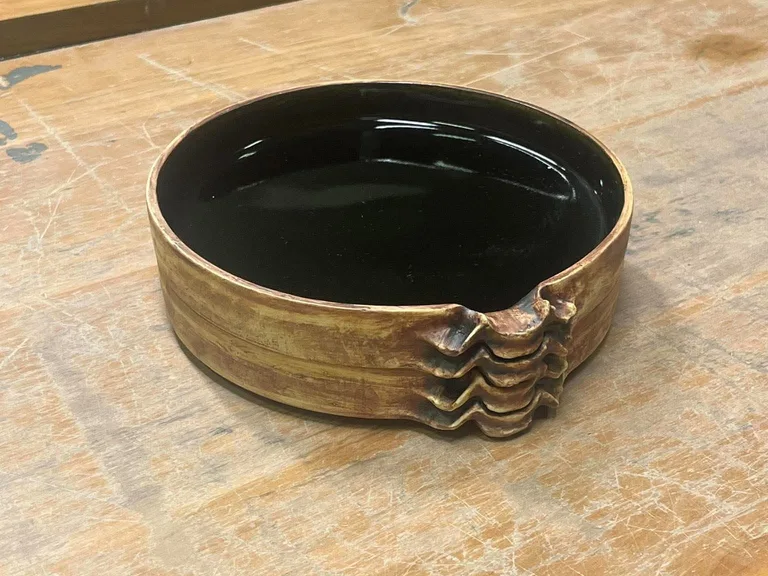 A low round plate, glazed black inside, with an ochre-brown outside and a spine-like relief, on a wooden workbench.