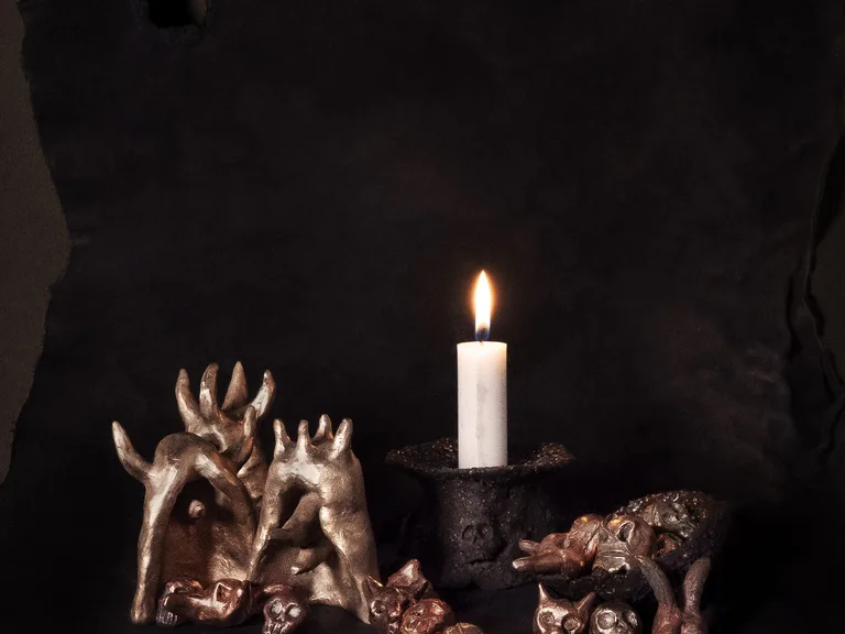 A lit candle in a dark, rough candleholder with a skull in relief, pale horned figures in a silver tone and small skulls and animal heads in copper and bronze tones, against a dark ground.
