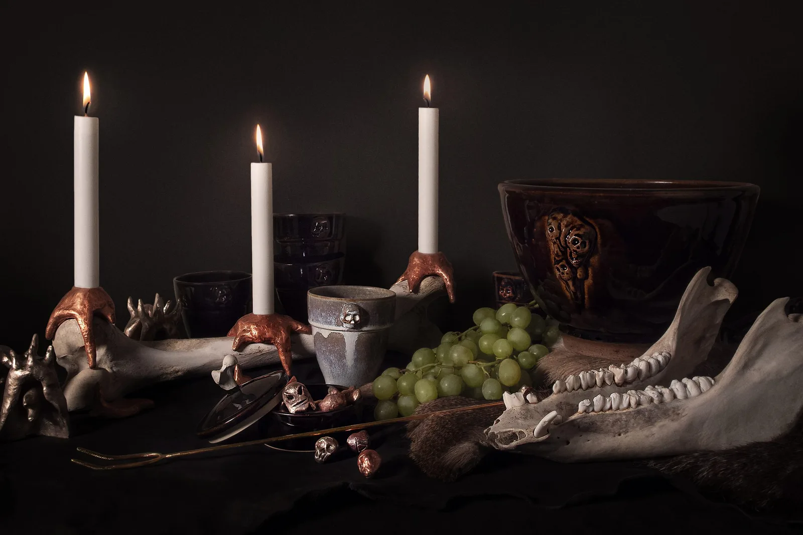 A table laid against a dark ground: three lit candles in copper-coloured candleholders gripping a large bone, ceramics with skulls in relief, grapes and an animal jaw on fur.