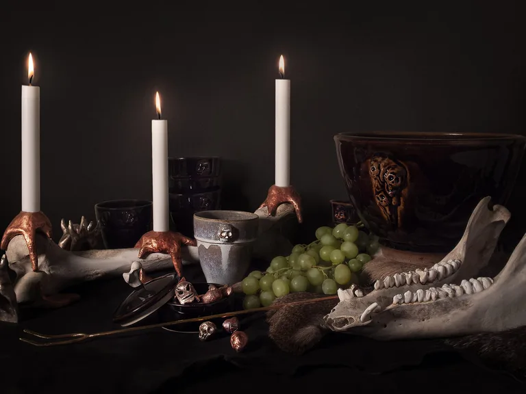 A table laid against a dark ground: three lit candles in copper-coloured candleholders gripping a large bone, ceramics with skulls in relief, grapes and an animal jaw on fur.