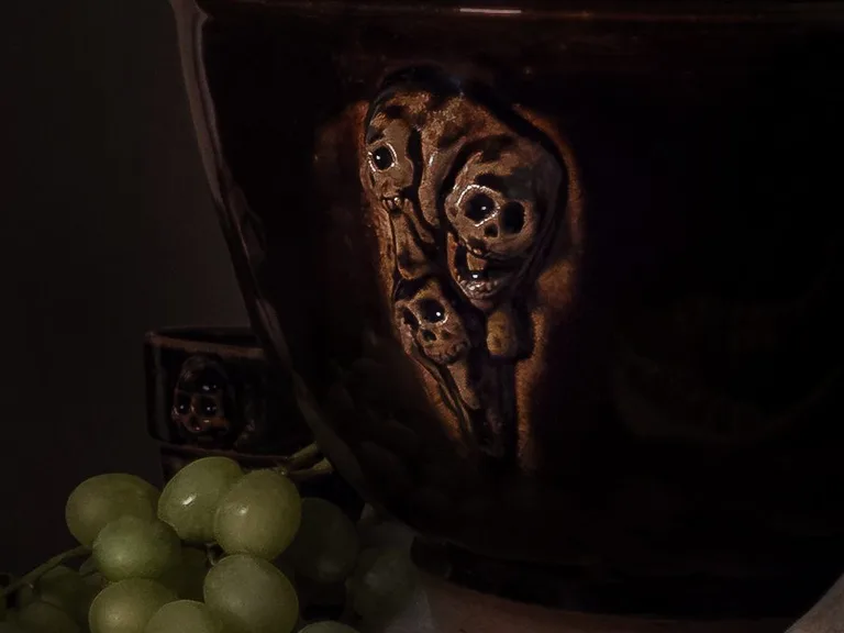 A dark brown glazed bowl with three skulls in relief, grapes and the teeth of an animal jaw on fur.