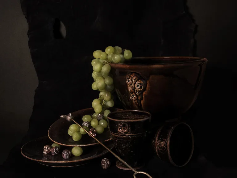 Two stacked dark-glazed plates with grapes and small skulls, a bowl with skulls in relief, a goblet with a skull, a toppled goblet and a long two-pronged fork.